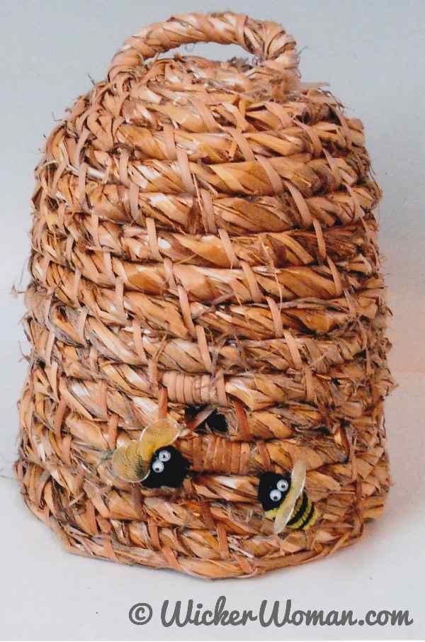 Rustic, straw, coiled bee skep with two little artificial bees at opening, woven by Cathryn Peters.